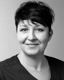Svartvit bild av en tandhygienist med ett vänligt leende, kort mörkt hår och iklädd en mörk arbetsrock.