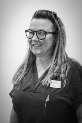En kvinna med långt, vågigt hår och glasögon, fotograferad i svartvitt. Hon ler och bär en svart arbetsuniform med en namnskylt, vilket indikerar att hon är en tandsköterska.