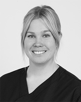 Svartvit porträttbild av en leende kvinna med uppsatt ljust hår, iförd en mörk topp, mot en ljus bakgrund.