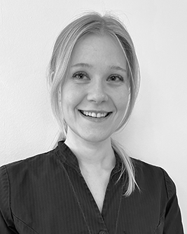 Svartvitt porträtt av en leende kvinna med ljust hår som är uppsatt. Hon bär en mörk krage eller arbetsuniform och står framför en ljus bakgrund