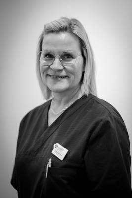 En leende tandsköterska med kort ljust hår, i svartvitt. Hon är iförd en svart arbetsuniform och har en namnskylt fäst på sin tröja.
