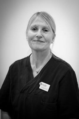 Porträtt av en kvinnlig tandhygienist med uppsatt ljust t hår och ett vänligt leende, iförd en mörk arbetsrock med en namnbricka, mot en ljus bakgrund i svartvitt.