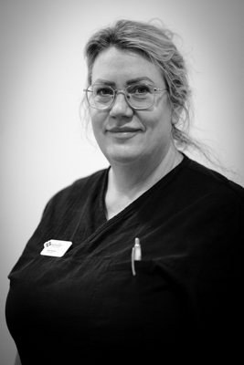Porträtt av en kvinnlig tandhygienist med glasögon, uppsatt ljust hår och ett vänligt leende, iförd en mörk arbetsrock med en namnbricka, mot en ljus bakgrund i svartvitt.