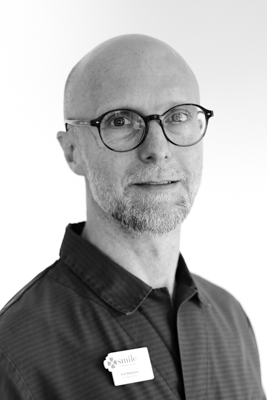 Porträtt av en manlig tandläkare med skallighet och glasögon, iförd en mörk arbetsrock med en namnbricka, som ger ett seriöst och professionellt intryck mot en ljus bakgrund i svartvitt.