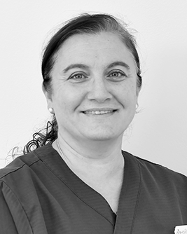 Porträtt av en kvinnlig tandhygienist med mörkt hår uppsatt i en elegant frisyr, ett leende på läpparna, klädd i en mörk arbetsdräkt, mot en enkel bakgrund.