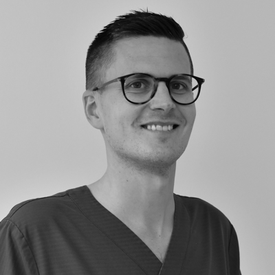 Porträtt av en manlig tandläkare med glasögon och mörkt hår, leende mot kameran, iförd en mörk arbetsdräkt med en namnbricka, mot en neutral bakgrund.