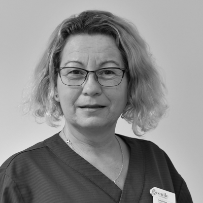 Porträtt av en kvinnlig tandhygienist med kort lockigt hår och glasögon, ett leende, klädd i en mörk arbetsdräkt, mot en neutral bakgrund.