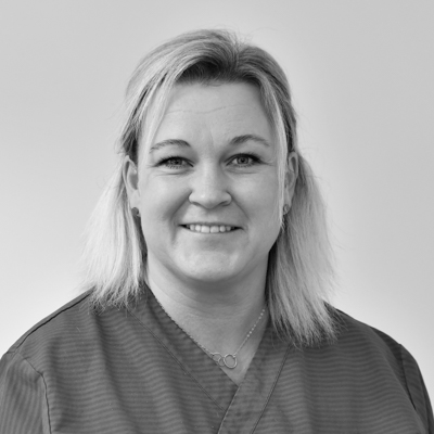 Porträtt av en kvinnlig tandhygienist med axellångt blont hår, leende mot kameran, klädd i en mörk arbetsdräkt, mot en neutral bakgrund.