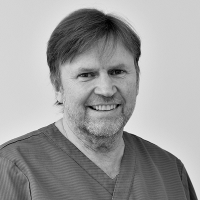 Porträtt av en manlig tandläkare med medellångt, mörkt hår och skägg, som ser direkt in i kameran med ett brett leende, klädd i en mörk arbetsdräkt.