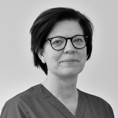 Porträtt av en kvinnlig tandsköterska med kort mörkt hår och stora glasögon, klädd i en mörk arbetsdräkt, med ett seriöst ansiktsuttryck, mot en neutral bakgrund.