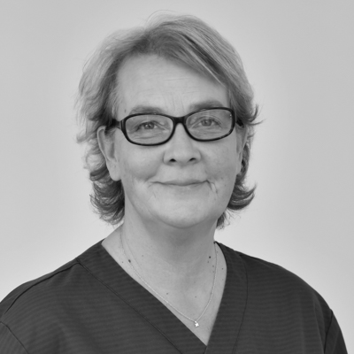 Porträtt av en kvinnlig tandhygienist med kortare hår och glasögon, ett vänligt uttryck, klädd i en mörk arbetsdräkt, mot en enkel bakgrund.