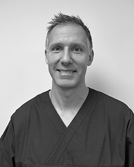 En tandläkare med poserar framför en ljus bakgrund, iklädd vårduniform och namnskylt, och ler mot kameran.