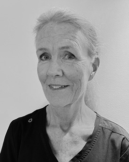 Svartvitt porträtt av en leende kvinna med ljust hår som är uppsatt. Hon bär en mörk krage eller arbetsuniform och står framför en ljus bakgrund