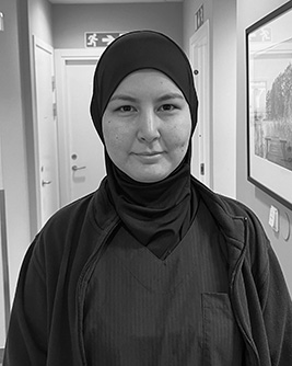 En person i hijab och arbetskläder står i en klinikliknande korridor och tittar rakt in i kameran.