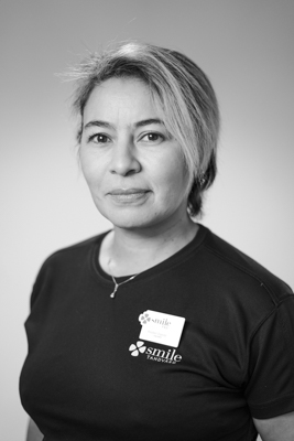 Porträtt av en kvinnlig tandhygienist med ett varmt leende, kort ljust hår, iförd en mörk arbetsrock, som ser vänligt mot kameran mot en suddig bakgrund i svartvitt.