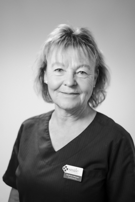 Porträtt av en kvinnlig tandsköterska med ljust kort hår, iförd en mörk arbetsrock, som ser direkt in i kameran med ett vänligt uttryck mot en ljus bakgrund i svartvitt.