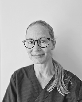 Porträtt av en leende oral kirurg med glasögon och långt hår uppsatt i hästsvans. Hon bär en mörk arbetsdräkt och står mot en ljus bakgrund.