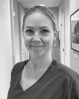 Svartvit bild av en tandsköterska med ett vänligt leende, uppsatt hår och klädd i en mörk arbetsrock. Bakgrunden visar en korridor med stängda dörrar, vilket indikerar en klinisk miljö.