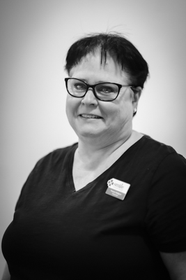 En kvinna med mörkt hår och glasögon, fotograferad i svartvitt, bär en mörk arbetsuniform med en namnskylt. Hon ser rakt in i kameran med ett professionellt uttryck.