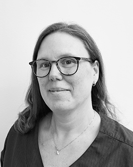 Porträtt av en kvinnlig team-service medarbetare inom tandvård med glasögon och axellångt hår, iförd en mörk arbetsrock, som ser direkt in i kameran med ett vänligt glatt uttryck mot en ljus bakgrund i svartvitt.
