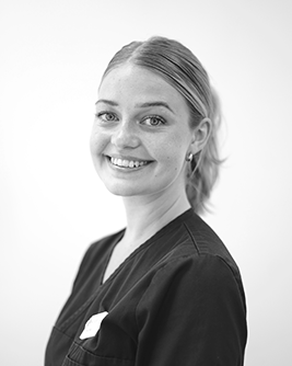 Glad tjej med blont, uppsatt hår som står i mörka arbetskläder, framför en vit vägg.