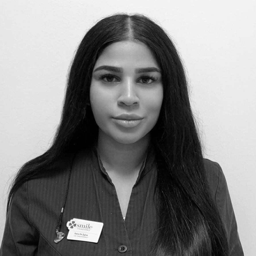 Porträtt av en kvinnlig tandhygienist med ett neutralt leende, långt mörkt hår, iförd en mörk arbetsrock, som ser vänligt mot kameran mot en suddig bakgrund i svartvitt.