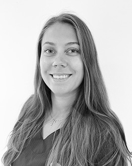 Porträtt av en kvinnlig medarbetare med långt hår, iförd en mörk arbetsrock, som ser direkt in i kameran med ett vänligt uttryck mot en ljus bakgrund i svartvitt.