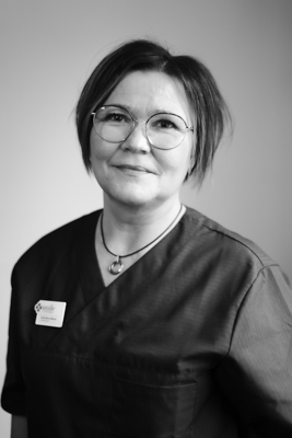 Porträtt av en tandhygienist med glasögon och mörk skrubbsuniform, hennes hår är stylat i en kort bob och hon bär ett namnbricka, ser vänlig och erfaren ut.