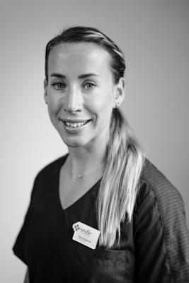 Porträtt av en leende tandhygienist i mörk skrubbsuniform, med långt ljust hår i en hästsvans och ett namnbricka, som ger ett energiskt och professionellt intryck.