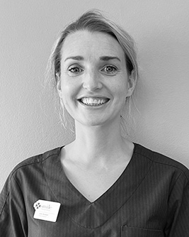 Svartvit bild av en leende kvinnlig tandhygienist som bär en arbetsuniform med namnbricka, stående mot en enkel bakgrund.