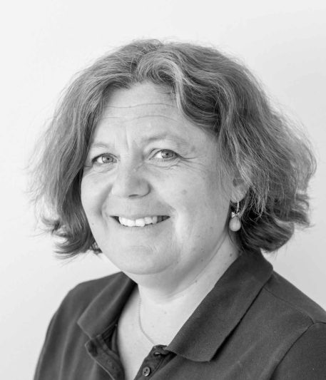 Porträtt av en kvinnlig tandhygienist med ett varmt leende, kort hår, iförd en mörk arbetsrock, som ser vänligt mot kameran mot en suddig bakgrund i svartvitt.