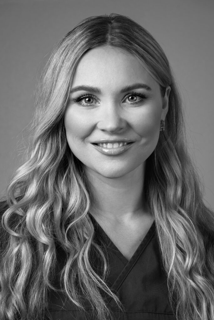 Porträtt av en kvinnlig team-service medarbetare inom tandvård med långt ljust hår, iförd en mörk arbetsrock, som ser direkt in i kameran med ett vänligt glatt uttryck mot en ljus bakgrund i svartvitt.