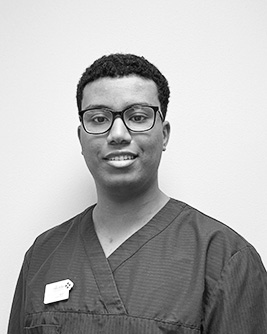 En tandhygienist med glasögon och kort lockigt hår poserar framför en ljus bakgrund, iklädd vårduniform och namnskylt, och ler mot kameran.