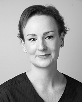 Porträtt av en kvinnlig tandläkare med kort mörkt hår och blå klinikkläder, som ser direkt in i kameran med ett självsäkert leende.