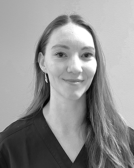 Porträtt av en ung kvinnlig tandhygienist med långt hår och ett mjukt leende, klädd i en mörk arbetsrock, som ser direkt in i kameran mot en suddig bakgrund i svartvitt.