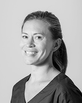 Porträtt av en kvinna med mörkt, uppsatt hår, iförd en mörk arbetsrock, som ser direkt in i kameran med ett vänligt uttryck mot en ljus bakgrund i svartvitt.