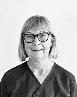 Porträtt av en kvinna med blont kort hår, iförd en mörk arbetsrock, som ser direkt in i kameran med ett vänligt uttryck mot en ljus bakgrund i svartvitt.