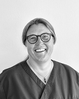 Porträtt av en kvinnlig Specialisttandläkare i Ortodonti med glasögon och uppsatt hår, iförd en mörk arbetsrock, som ser direkt in i kameran med ett vänligt uttryck mot en ljus bakgrund i svartvitt.