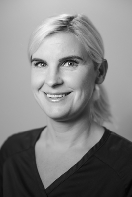 Porträtt av en kvinnlig tandläkare med uppsatt ljust hår och ett vänligt leende, iförd en mörk arbetsrock, mot en ljus bakgrund i svartvitt.