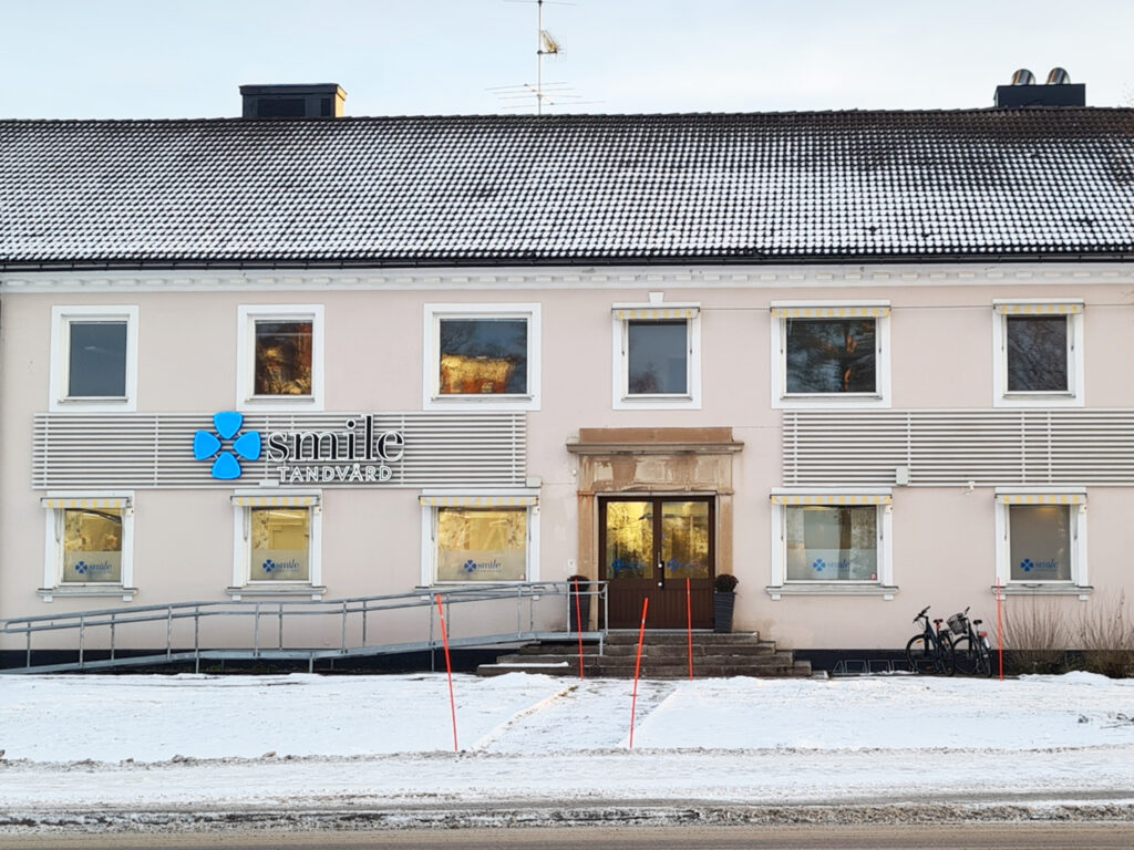 En tvåvånings byggnad målad i ljusa färger med skylten Smile Tandvård syns tydligt placerad på fasaden, belägen i en vintermiljö. Kliniken ser ut att vara väl underhållen, med en traditionell arkitektur som passar för medicinska tjänster, och tjänar som en tandläkarpraktik i Boden.
