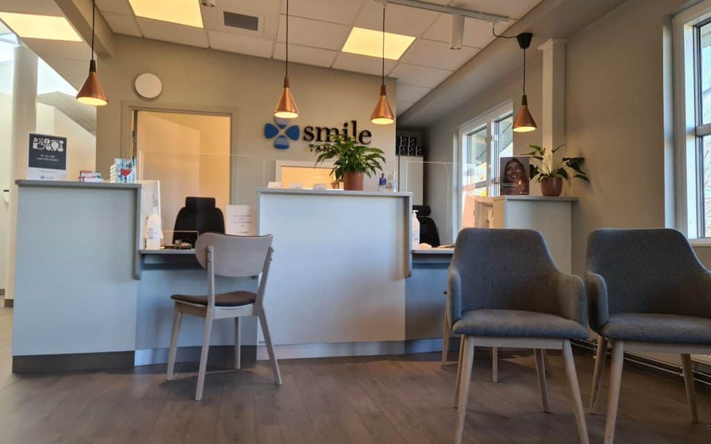 Bilden visar ett väntområde i Smile Tandvård Sandvikens tandläkarklinik. Det finns fyra stolar i blått tyg för patienter att sitta på medan de väntar. Ett litet bord står mellan två av stolarna. I bakgrunden syns receptionsdisk där en anställd kan ta emot patienter. Över disken hänger en skylt med texten "smile". Belysningen kommer från pendellampor ovanför disken samt från fönster till vänster i bilden som inte syns. Det finns även inomhusväxter som bidrar till en välkomnande atmosfär. Designen är enkel och modernt avskalad.