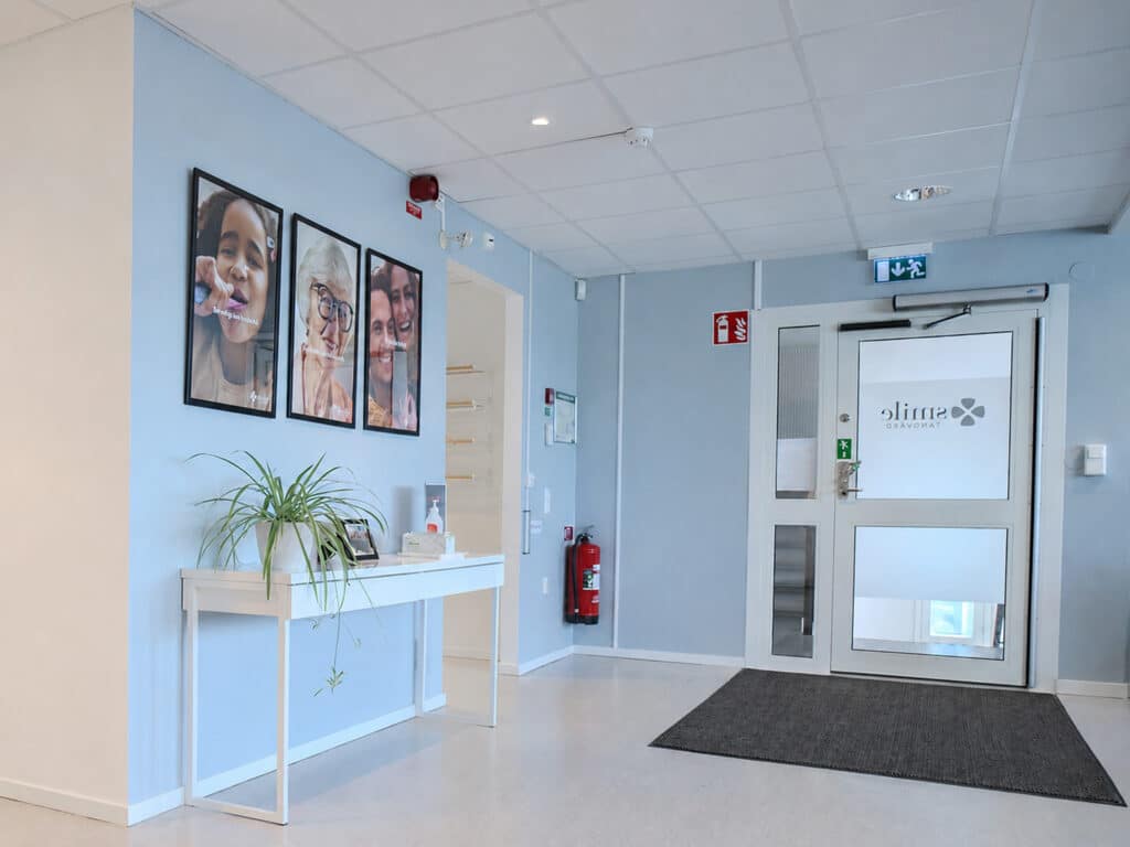 Entré och väntrumsområde på Smile Tandvård Jönköping City med ljusblå väggar, informationsbord med grön växt, patientbilder på väggen och glasad entrédörr.