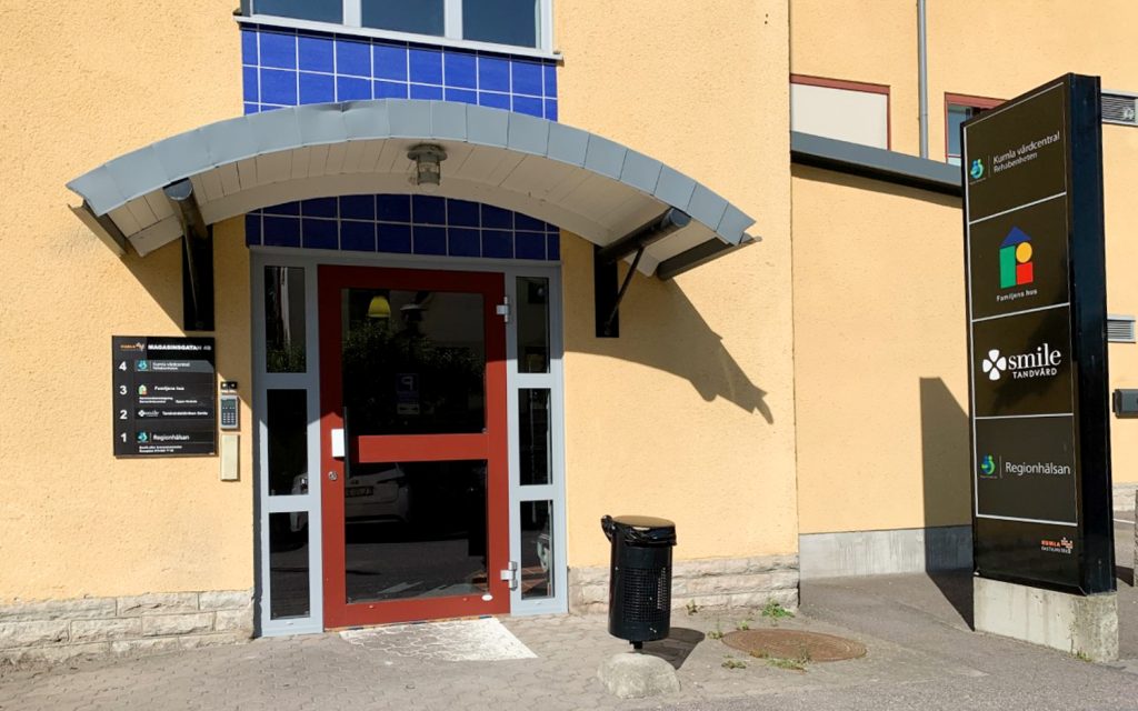 Exteriörbild av Smile Tandvård i Kumla, som visar huvudingången med en röd dörr och en blå och vit baldakin. Tandvårdsklinikens skylt och övriga företagsskyltar visas på en stolpe bredvid ingången, som tydligt markerar kliniken i ett kommersiellt område.