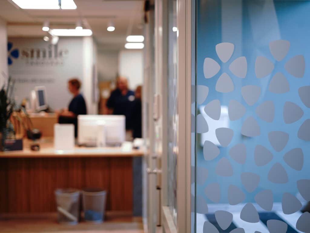 Ingången till Smile Tandvård i Norrköping med en närbild på en glasdörr dekorerad med klinikens logotyp och mönster. I bakgrunden syns receptionen med en receptionsdisk i trä, en dator och krukväxter. Receptionisten och ytterligare personal arbetar bakom disken. Väggarna är vita och dekorerade med klinikens namn och logotyp, vilket ger en modern och professionell känsla.
