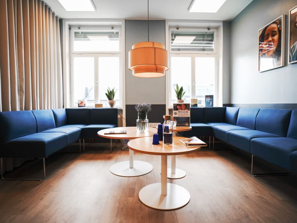 En väntrumsmiljö på Smile Tandvård i Norrköping med moderna blå soffor, runda träbord och växter som skapar en inbjudande atmosfär. Taklampan i mitten ger ett varmt ljus och fönstren släpper in naturligt dagsljus, vilket ger rummet en ljus och luftig känsla. På väggarna hänger konstverk som bidrar till en avslappnad och trevlig stämning.