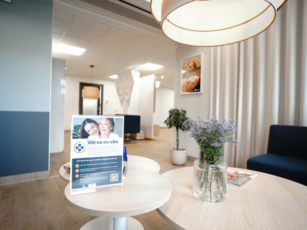 Interiör av väntrummet på Smile Tandvård i Norrköping med moderna blå soffor, vita runda bord och en vas med blommor på bordet. En broschyr om tandvårdstjänster står på ett av borden, och taklampor ger ett varmt ljus över rummet. Bakgrunden visar fler sittplatser och en ljus och välkomnande atmosfär med neutrala färger och väggdekorationer.