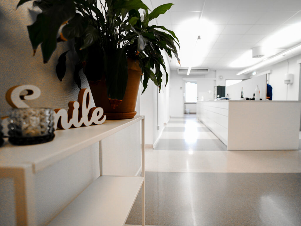 Interiörbild från Smile-kliniken i Boden, som visar en ljus och modern korridor med vita väggar och golv. I förgrunden står en hylla med en dekoration som stavas 'Smile' och en krukväxt. Längre in i bilden syns en reception eller arbetsyta med vita bänkar och utrustning. Rummet är upplyst med taklampor och skapar en ren och inbjudande atmosfär.