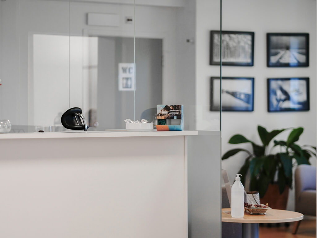 Receptionen på Smile-kliniken i Boden, med en modern vit disk som har en glasavskärmning. På disken står en bukett konstgjorda blommor och ett kortställ. I bakgrunden syns ett väntrum med växter och väggar dekorerade med inramade fotografier. Ett litet bord med handsprit och broschyrer är placerat framför väntrumsstolarna.