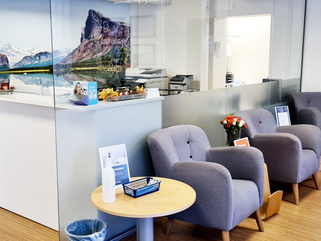 Väntrummet på Smile Tandvård i Boden med bekväma grå stolar, ett runt bord med läsmaterial, och en receptionsdisk. Bakom disken syns en stor väggmålning som framställer en naturskön vy, vilket skapar en lugn och välkomnande atmosfär.