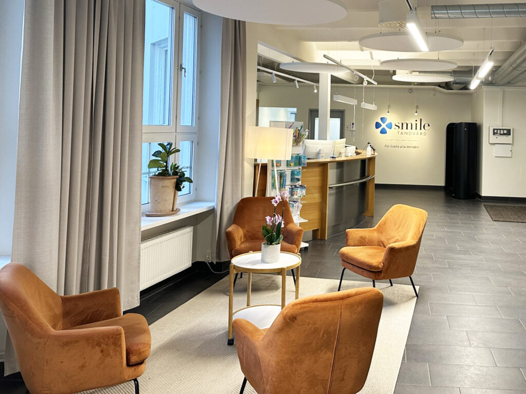 En modern loungehörna med en brun sammetsfåtölj, ett runt sidobord med två värmeljus, en vit golvlampa och en krukväxt placerad i fönstret framför ljusgrå gardiner.